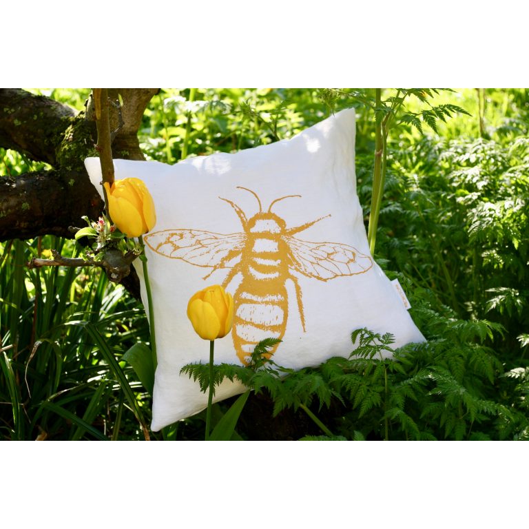 Frohstoff Kissen aus Leinen mit Bienen Motiv in geld zwischen Pflanzen liegend und an einen Ast gelehnt, davor gelbe Blumen