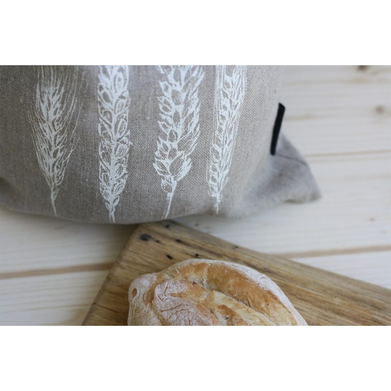 Frohstoff Nahaufnahme vom Brotbeutel mit Ähren Motiv in Perlmutt auf einem weißen Holztisch. Auf dem Tisch liegt ein Holzbrett mit einem Brot drauf