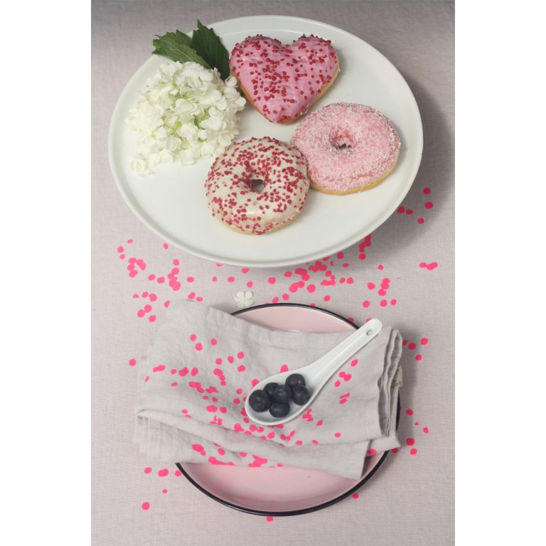 Frohstoff Servietten aus Leinen und Tischläufer aus Leinen mit Blütenblätter motiv in pink. Die Servietten sind auf einem Teller zusammengelegt darauf befindet sich ein weißer Porzellan Löffel mit Beeren. Auf der oberen Seite des Bildes steht ein Teller mit Donuts