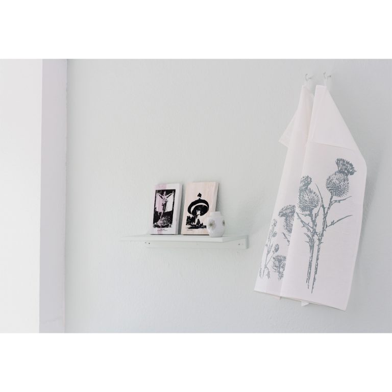 Frohstoff 2 Geschirrtücher mit Distel Motiv nebeneinander an einer Wand aufgehängt, links daneben hängt ein dekoriertes Regal