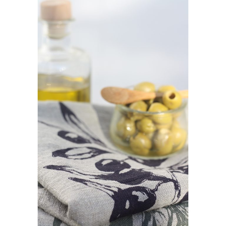 Frohstoff Geschirrtuch aus Leinen Natur mir Oliven Motiv leicht versetzt aufeinander zusammengelegt dekoriert mit einer Flasche mit Olivenöl und einer Schale gefüllt mit Oliven und einem Holzlöffel