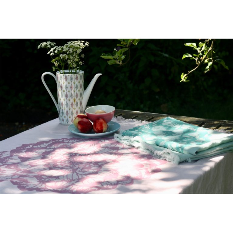 Frohstoff Plaid aus Leinen rosa und Minze mit dem Dahlie Motiv dekoriert auf einem Holztisch dekoriert mit einer gemusterten Gießkanne in der sich ein Blumenstrauß befindet, einem Teller mit einer Schüssel und Äpfeln drauf