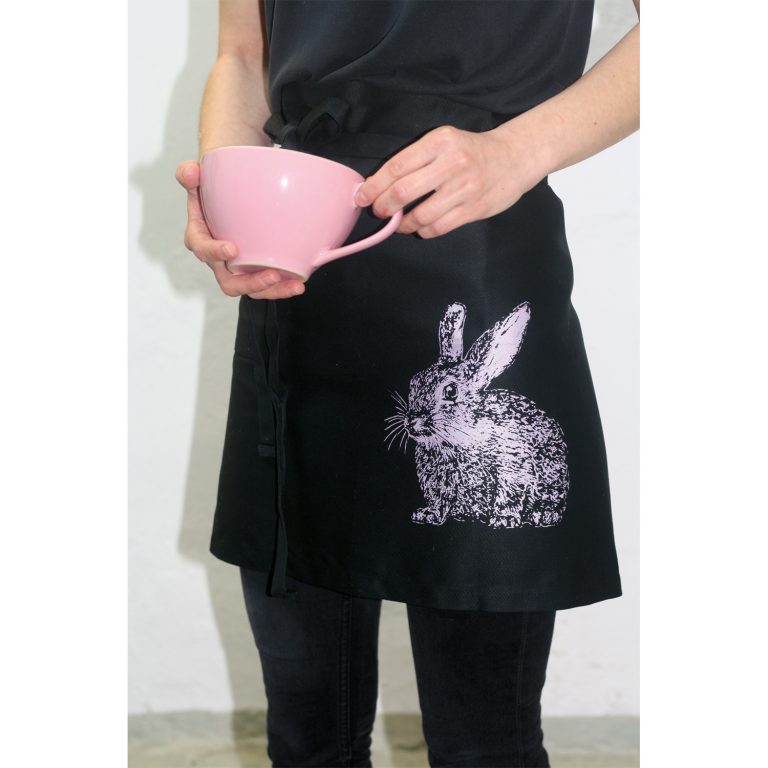 Frohstoff Vorbinderschürze schwarz mit Wildkaninchen Motiv in Rosa an einer Frau mit einer Rosanen Tasse in der Hand
