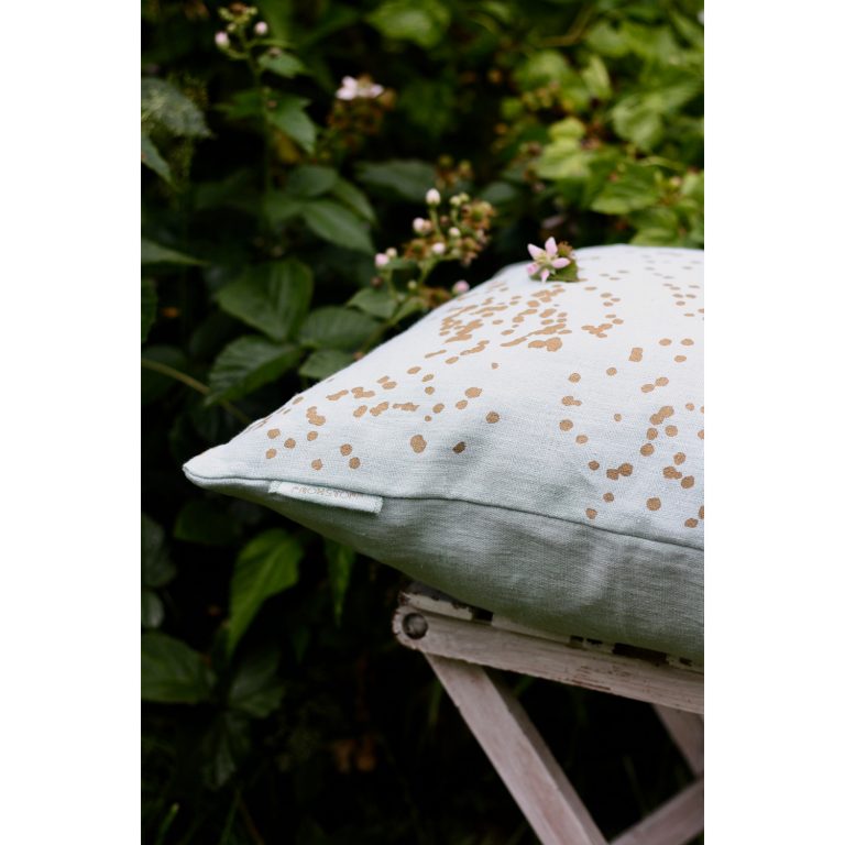 Frohstoff Kissen aus Leinen Grau mit Blüten Motiv in messing auf einem hölzernen Hocker vor einer Blätterwand
