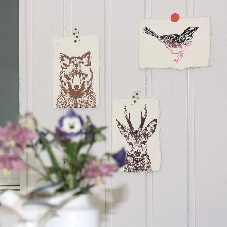 Frohstoff 3 Kunstdrucke mit Fuchs, Hirsch und Spatz Motiv an einer weißen Holzwand aufgehängt mit einem Blumenstrauß im Vordergrund