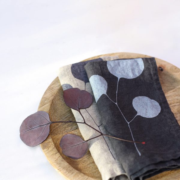 Frohstoff Serviette aus Leinen Natur und Anthrazit mit Eukalyptus-Motiv auf einem Holzteller mit einem Eukalyptus Blatt