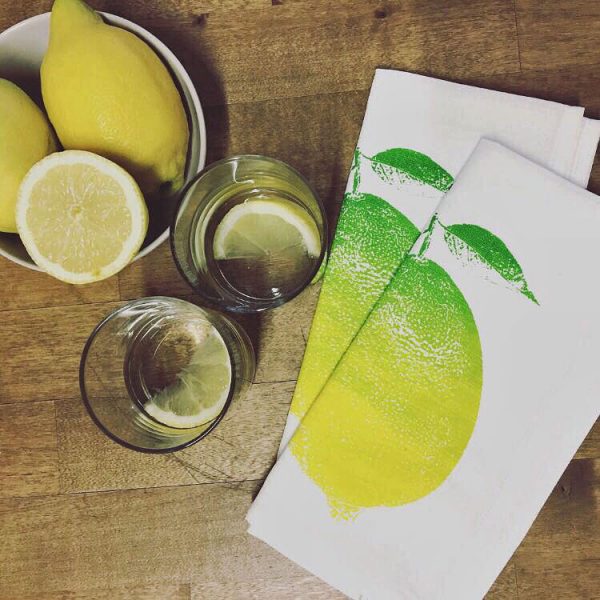 Frohstoff Serviette Zitrone grün-gelber farbverlauf zusammengelegt auf einem Holztisch daneben 2 Gläser mit Zitronenscheiben und eine Schüssel mit Zitronen von Oben fotografiert