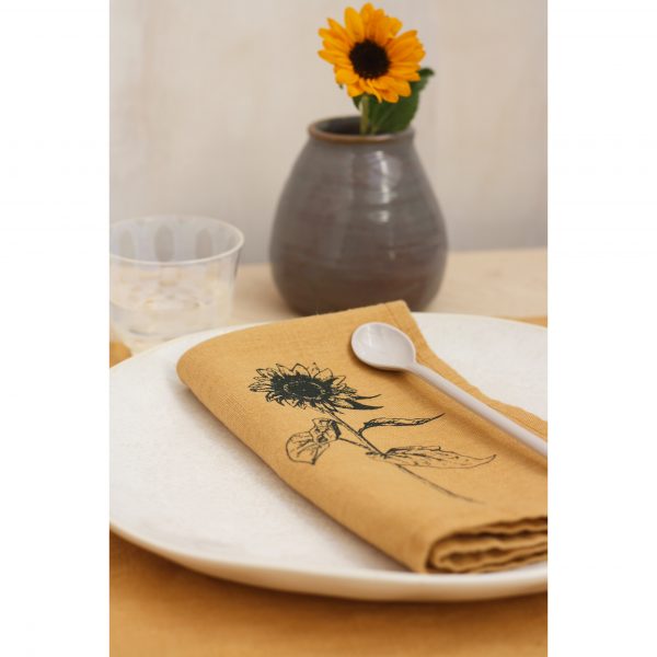 Frohstoff Serviette und Tischläufer in Honiggelb mit Sonnenblumen-Motiv zusammen dekoriert. Eine Sonnenblume in einer Phase steht im Hintergrund