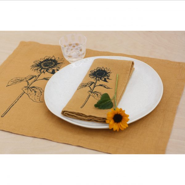 Frohstoff Tischset und Serviette in Honiggelb mit Sonnenblumen-Motiv zusammen dekoriert, Die Serviette liegt auf einem Teller zusammen mit einer kleinen Sonnenblume auf der obigen linken Seite steht ein Glas