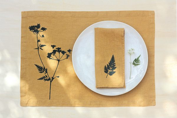 Frohstoff Tischset aus Leinen Honiggelb mit Wiesenkerbel-Motiv und Serviette aus Leinen Honiggelb mit Blattmotiv. Die Serviette ist zusammengelegt auf einem Teller angerichtet